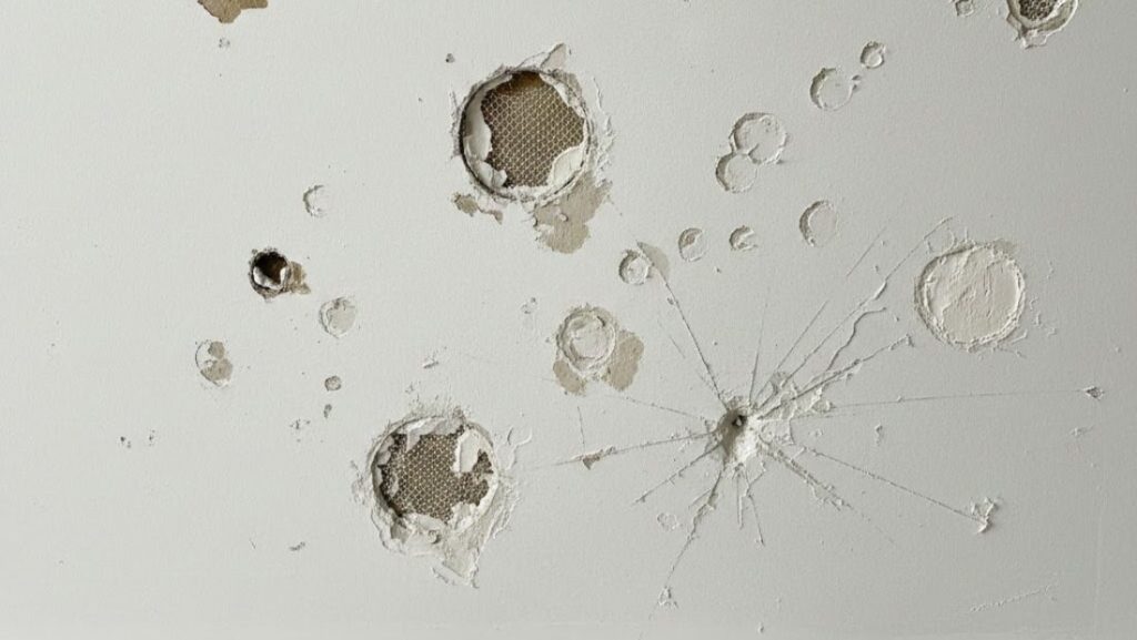Multiple drywall holes and dents on a white wall surface, including small punctures, larger damaged areas exposing mesh, and visible impact cracks radiating outward.