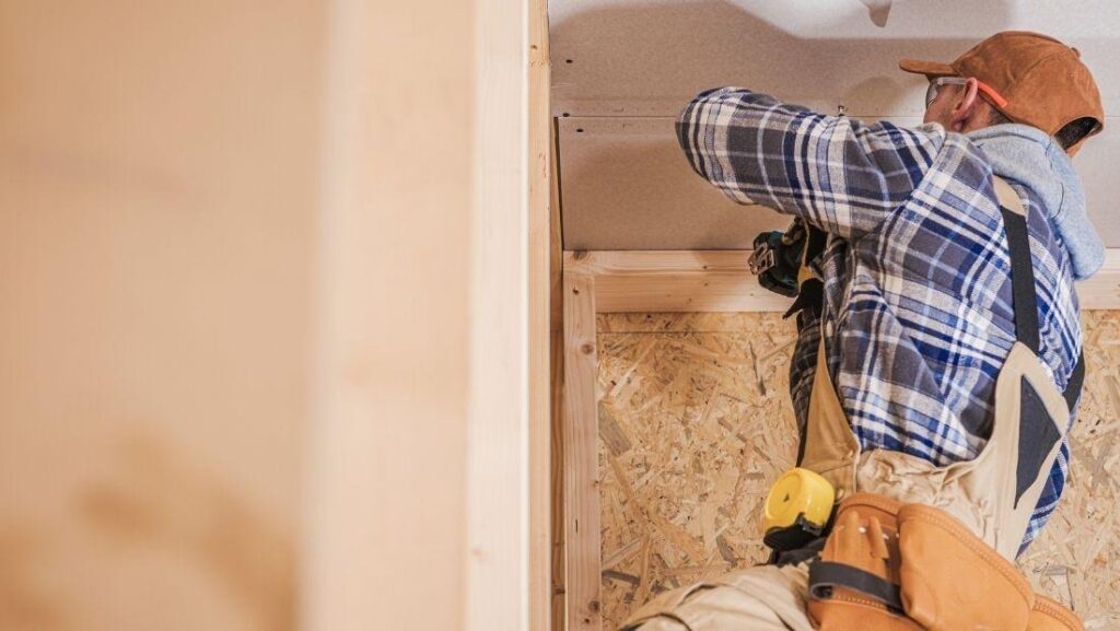 A worker patching drywall, wearing a plaid shirt and tool belt, using a drill to secure drywall panels in a wooden frame. This process helps repair holes in the wall for a smooth finish.
