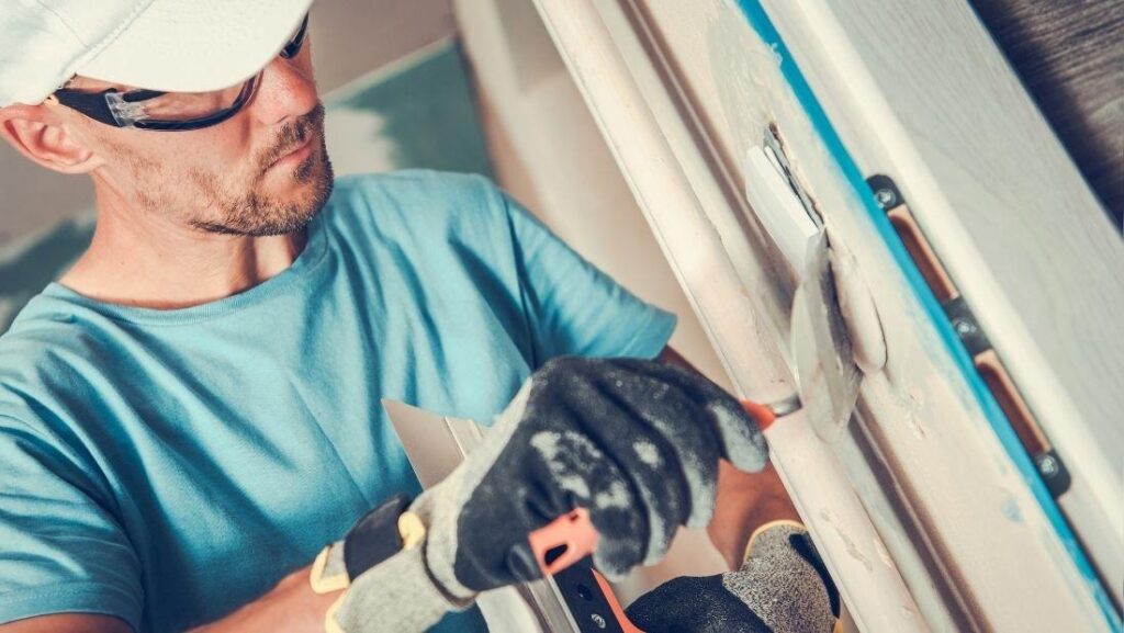 A worker using a drywall knife to patch drywall, filling in a hole with joint compound. The worker is wearing gloves and safety glasses while carefully smoothing the surface.