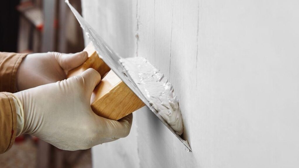 Repairing cracked plaster walls using a putty knife to apply fresh plaster smoothly over surface damage on an interior wall.