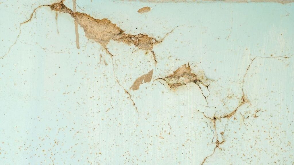 Cracked plaster walls with peeling paint, visible surface damage, and spreading fissures across a light-colored interior wall, showing signs of wear and structural deterioration.