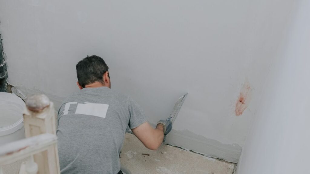 A worker performing wall repairs, using a trowel to smooth out a patched area on a white wall.