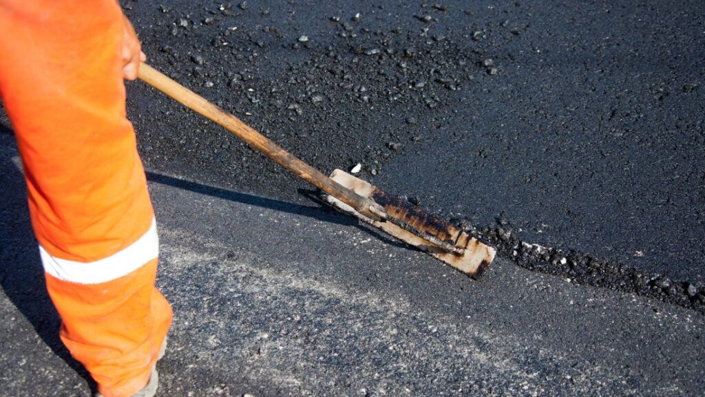 Worker demonstrating how to repair an asphalt parking lot by cleaning and preparing a damaged crack in the pavement using a tool